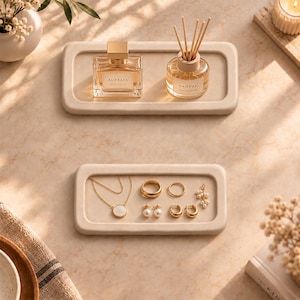 May include: Two rectangular beige trays with rounded corners. The top tray holds a glass perfume bottle labeled Aurelia and a reed diffuser labeled Santal. The bottom tray contains a gold necklace, a gold ring, and several pairs of earrings.