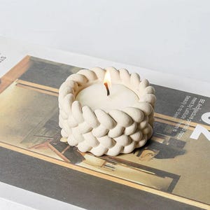 May include: A white ceramic candle holder with a braided design. The holder has a small, white candle inside.