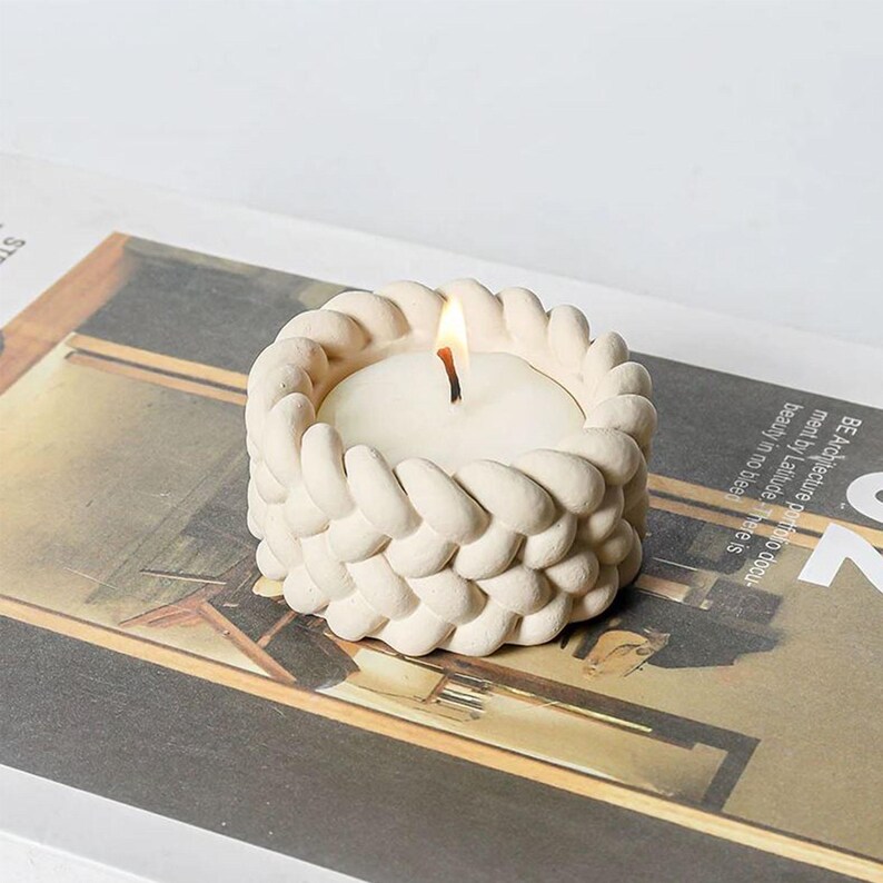 May include: A white ceramic candle holder with a braided design. The holder has a small, white candle inside.