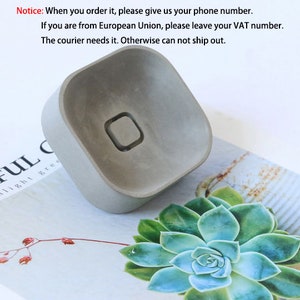 May include: A gray concrete square planter with a small hole in the center. The planter is sitting on a white surface with a green succulent plant in the background.