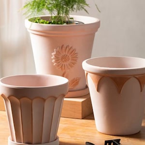 Pode incluir: Três vasos de flores de terracota na cor terracota. O vaso maior tem um desenho de girassol em relevo. Os outros dois vasos têm uma borda festonada.