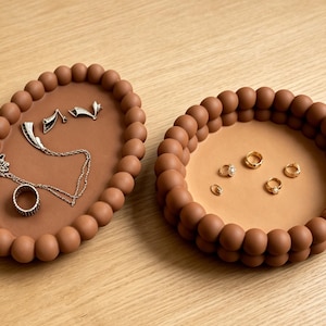 May include: An oval tray and a circular tray, both featuring a raised border of matte brown spheres. The oval tray holds a silver chain necklace and a ring, while the circular tray contains five small gold rings on a light tan base.