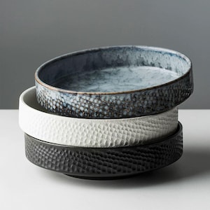 May include: Three ceramic bowls stacked on top of each other. The top bowl is blue with a speckled glaze, the middle bowl is white with a textured surface, and the bottom bowl is black with a textured surface.