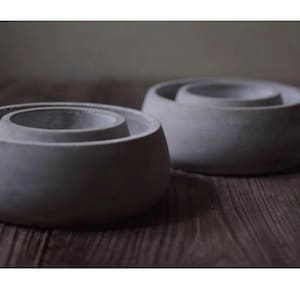 以下が含まれることがあります： 円形のデザインの2つの灰色のコンクリートボウル。 各ボウルには、中央に隆起したリングがあり、層状の効果を生み出しています。 ボウルは木製の表面に置かれ、ミニマリストでモダンなデザインに焦点を当てています。