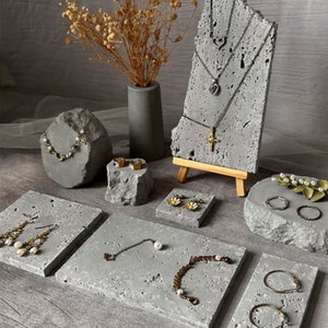 Pode incluir: Um conjunto de expositores de joias de concreto cinza. Os expositores são de várias formas e tamanhos, incluindo um expositor retangular alto com um cavalete de madeira, um expositor redondo e vários expositores quadrados. Os expositores são perfeitos para exibir colares, pulseiras, brincos e anéis.