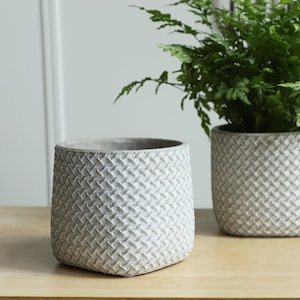 May include: Two grey concrete planters with a woven pattern. The planters are empty and sitting on a wooden surface.