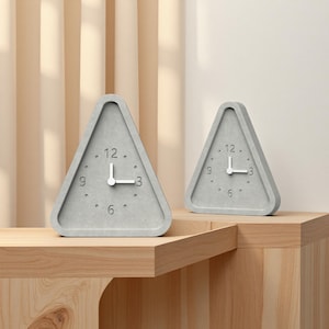 Puede incluir: Dos relojes triangulares grises con manecillas blancas y números negros. Los relojes están colocados sobre una superficie de madera. Los relojes tienen un diseño minimalista y son un elemento de decoración moderno.