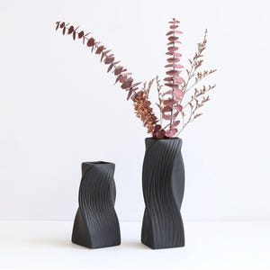 May include: Two black ceramic vases with a twisted design. The taller vase holds dried eucalyptus and other dried flowers. The vases are on a white surface. Home decor.