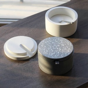 Puede incluir: Tres recipientes redondos de hormigón con tapas. Dos son blancos y uno es gris con motas blancas. Las tapas tienen una pequeña abertura en el centro.