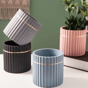 Puede incluir: Conjunto de macetas cilíndricas en negro, gris y azul. Cada maceta presenta detalles de estrías verticales y una banda dorada. Una maceta contiene una planta verde, mientras que otras están vacías.