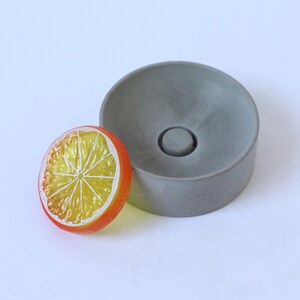 May include: A gray concrete dish with a round, slightly raised center. The dish is about 4 inches in diameter. A yellow and orange plastic citrus slice is next to the dish.