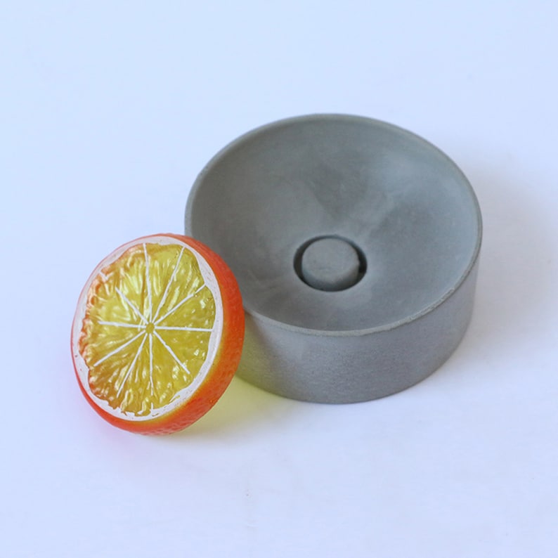 May include: A gray concrete dish with a round, slightly raised center. The dish is about 4 inches in diameter. A yellow and orange plastic citrus slice is next to the dish.