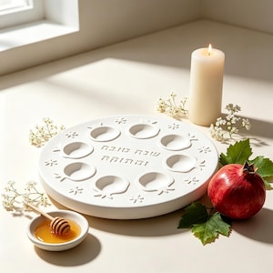 May include: A white circular ceramic tray with eight pomegranate-shaped indentations and Hebrew text in the center. The tray is accompanied by a small white bowl containing honey and a wooden dipper, a lit pillar candle, and a fresh pomegranate.
