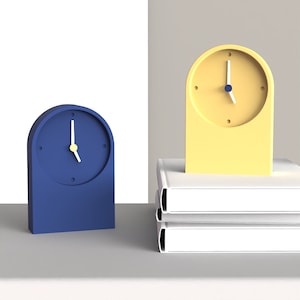 Puede incluir: Dos despertadores analógicos, uno azul y otro amarillo, están sobre una superficie blanca. El despertador amarillo está encima de una pila de tres libros blancos.