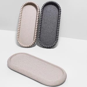Può includere: Tre vassoi decorativi in tonalità neutre. Due sono ovali con bordi perlati, uno beige e l'altro grigio scuro. Il terzo vassoio è ovale con una superficie a coste, in color beige chiaro. Misure in centimetri.