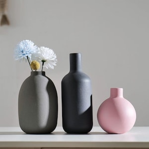 May include: Three ceramic vases in a row. The first is grey and holds light blue and white flowers. The second is black and bottle-shaped. The third is a small, round, pink vase. All vases are on a white surface.