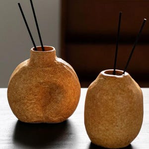May include: Two textured, tan-colored ceramic vases with a rough, stone-like finish. Each vase has a narrow neck and contains two thin, black reed diffuser sticks protruding from the opening. The vases are rounded and have a matte, earthy appearance.