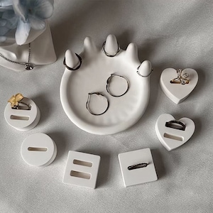 May include: White ceramic ring holder with multiple compartments for rings and earrings. The holder is shaped like a hand with five fingers. There are also two heart-shaped holders and two square holders.
