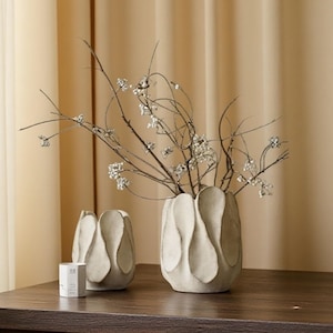 May include: Two off-white, textured vases with a petal design. The larger vase contains branches with small, white, beaded accents. A small, white, hexagonal box is to the left. The vases are on a dark wooden surface.