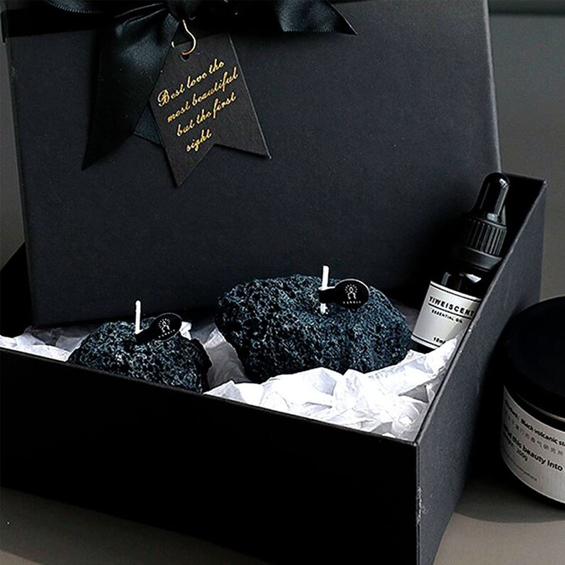 May include: A black gift box with a black ribbon and a tag that says "Don't love the most beautiful but the first sight". Inside the box are two black volcanic rock candles and a small bottle of essential oil.