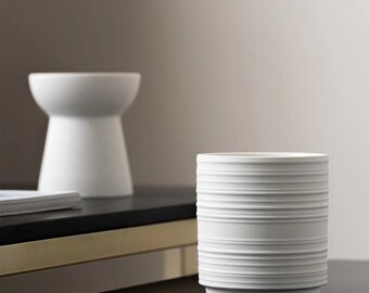 May include: Two white ceramic decorative objects. One is a cylindrical vase with horizontal ridged detailing. The other is a small, pedestal-style bowl. Both objects are on a dark surface with a neutral background.