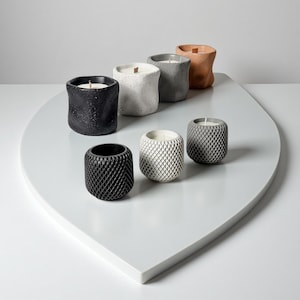 May include: An assortment of handcrafted candles in various colors and textures. The candles are in cylindrical containers, with colors including black, white, gray, and terracotta. Some have textured surfaces, while others have a diamond pattern. The candles are arranged on a white, leaf-shaped surface.