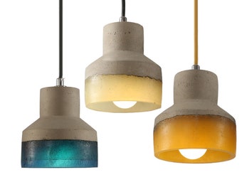 May include: Three pendant lights featuring cylindrical concrete tops and translucent lower sections in blue, white, and amber. Each fixture hangs from a cord, with the blue and white lights on black cords and the amber light on a yellow cord.
