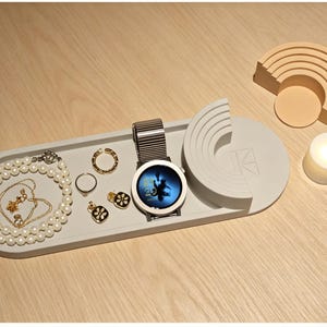 Puede incluir: Una bandeja ovalada gris contiene joyas, incluyendo un collar de perlas, anillos de oro y pendientes. Un reloj plateado con pantalla negra está junto a las joyas. También hay un arco decorativo y una vela encendida.