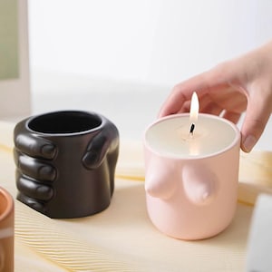 Puede incluir: Dos velas de cerámica, una con forma de mano y la otra con forma de busto. La vela con forma de busto es rosa y tiene una mecha encendida.