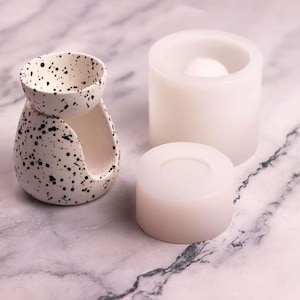 Könnte beinhalten: Ein weißer Keramik-Ölbrenner mit schwarzem Sprenkelmuster und ein passendes weißes Silikonform-Set, bestehend aus einem zylindrischen Sockel und einem Oberteil, das zum Gießen der Ölbrennerform in einheitlicher Größe dient.