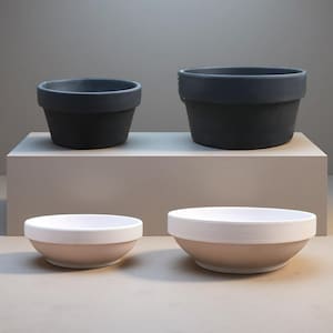 May include: Two dark gray ceramic plant pots of different sizes sit on a white platform. Below, two bowls with a white interior and a light pink exterior are displayed. The items are arranged on a neutral background.