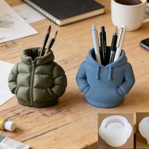 May include: Two novelty pen holders in the shape of clothing items are on a wooden desk. One is olive green and resembles a puffer jacket with a zip, and the other is blue and looks like a hoodie. Both contain pens and brushes.