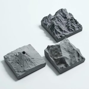 May include: Three gray, square-shaped mountain sculptures with detailed textures. One features a volcano with a central crater. The other two depict rugged mountain ranges, all set against a white background. These are likely decorative items.