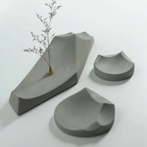 Może przedstawiać: Trzy szare betonowe tace o unikalnym, organicznym i fakturowanym designie. Największa taca jest długa i wąska, podczas gdy pozostałe dwie są mniejsze i bardziej okrągłe. Tace idealnie nadają się do wyeksponowania małych przedmiotów lub roślin.