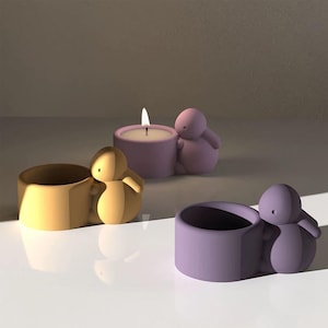 Op de afbeelding: Drie kaarshouders in de vorm van een persoon die een kleinere persoon vasthoudt. De kaarshouders zijn in geel, paars en donkerpaars. De middelste kaarshouder heeft een brandende kaars erin.