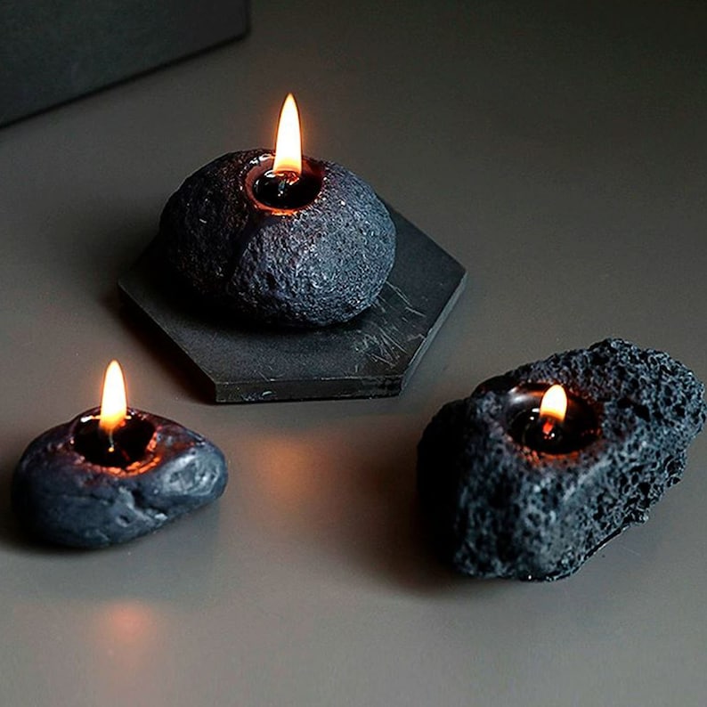 May include: Three black candle holders shaped like volcanic rocks with a small candle in each. The candles are lit and the flames are visible.