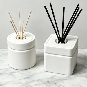 May include: Two white ceramic reed diffusers. One is a cylindrical vessel with light beige reeds, and the other is a square vessel with black reeds. Both feature a silver-toned metal collar at the opening where the reeds are inserted.