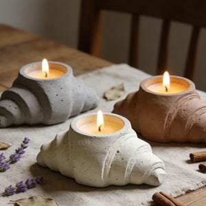 Puede incluir: Tres candelabros con forma de croissant en gris, blanco y marrón, cada uno con una vela encendida. Las velas están sobre un paño de lino de color neutro, con lavanda y palitos de canela cerca. Los candelabros son de un material texturizado.