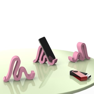 Puede incluir: Tres soportes de teléfono rosa con forma de línea ondulada. Un soporte sostiene un teléfono inteligente negro. Una unidad USB roja y negra está sobre una superficie verde claro.
