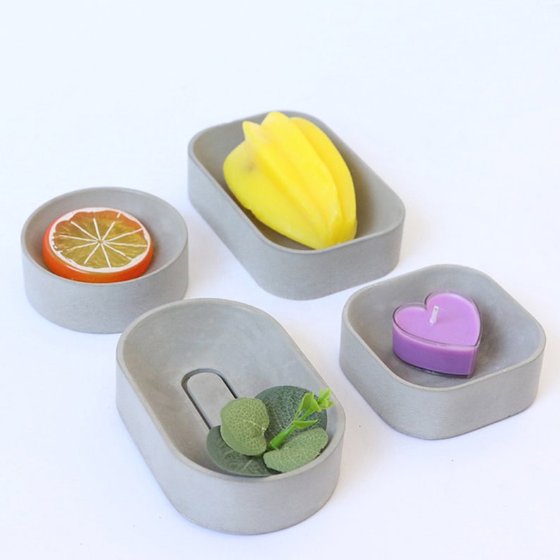May include: Four small gray concrete trays with different shapes. One tray holds a yellow starfruit, another holds a purple heart-shaped candle, another holds a slice of orange, and the last tray holds a small sprig of green leaves.
