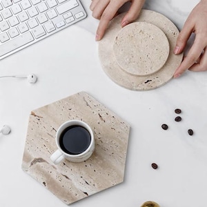 May include: A hexagonal beige stone coaster with a white coffee cup filled with dark liquid. A round stone coaster and a smaller round coaster are also present. Coffee beans and a keyboard are visible.
