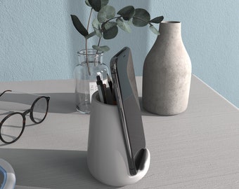 以下が含まれることがあります： 白いセラミック製の電話とペンのホルダー。黒いスマートフォンとペンがいくつか入っています。ホルダーは薄い灰色の表面に置かれており、背景には緑の植物の入った花瓶と灰色の花瓶があります。