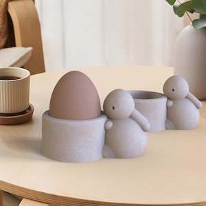 Puede incluir: Un soporte para huevos de cerámica blanca con forma de tres conejos. El soporte tiene un huevo marrón en uno de los nidos de conejo.