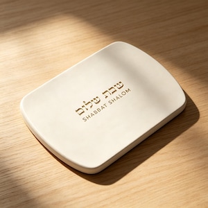 May include: A rectangular off-white tray with rounded corners featuring the Hebrew text Shabbat Shalom and the English text SHABBAT SHALOM embossed in gold lettering on the center of the smooth, matte surface of the tray.