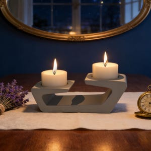 May include: A light gray concrete candle holder with a curved, asymmetrical design holding two lit white tea light candles. To the left is a small bundle of dried purple lavender, and to the right sits a gold-toned vintage pocket watch.