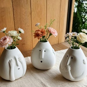Puede incluir: Tres jarrones de cerámica blanca con diseños de caras únicos, cada uno con un pequeño ramo de flores. Los jarrones tienen diferentes expresiones: uno se cubre los ojos, otro tiene una nariz larga y otro se sujeta la barbilla. Las flores incluyen flores rosas, blancas y naranjas.