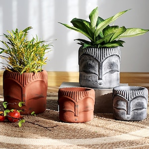 May include: Four decorative terracotta and gray planters, each with a stylized face design. Two planters are filled with green and yellow plants. The planters vary in size and are arranged on a woven mat.