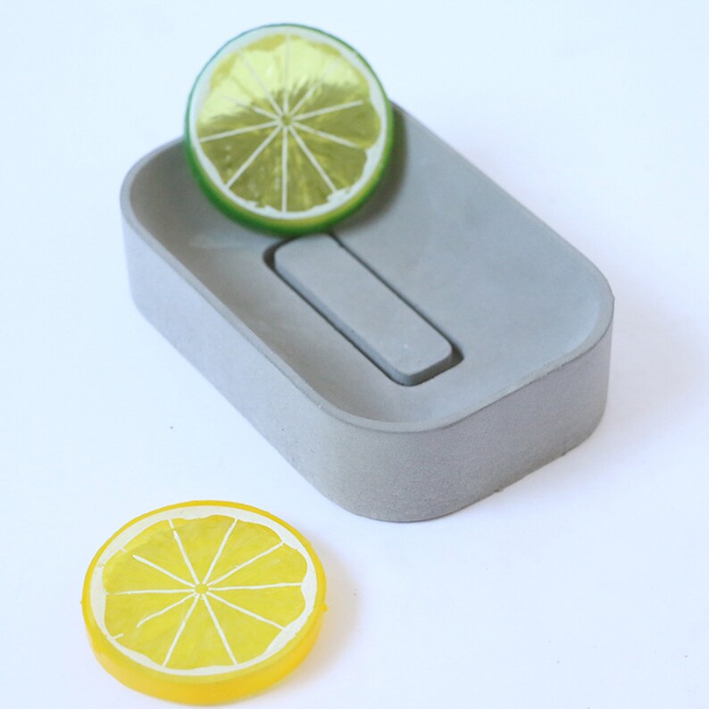 May include: A gray concrete rectangular dish with a small rectangular indentation in the center. A yellow lemon slice is on the left side of the dish and a green lemon slice is on the right side of the dish.