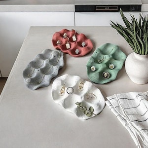 May include: Four decorative concrete trays in different colors, each with a wavy, organic shape. The trays are designed to hold jewelry and other small items. The trays are on a light gray surface.