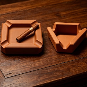 May include: A square terracotta-colored ashtray with four notches and a triangular terracotta-colored ashtray with three notches. A brown cigar with a gold band rests diagonally across the square ashtray, which features a smooth matte finish.